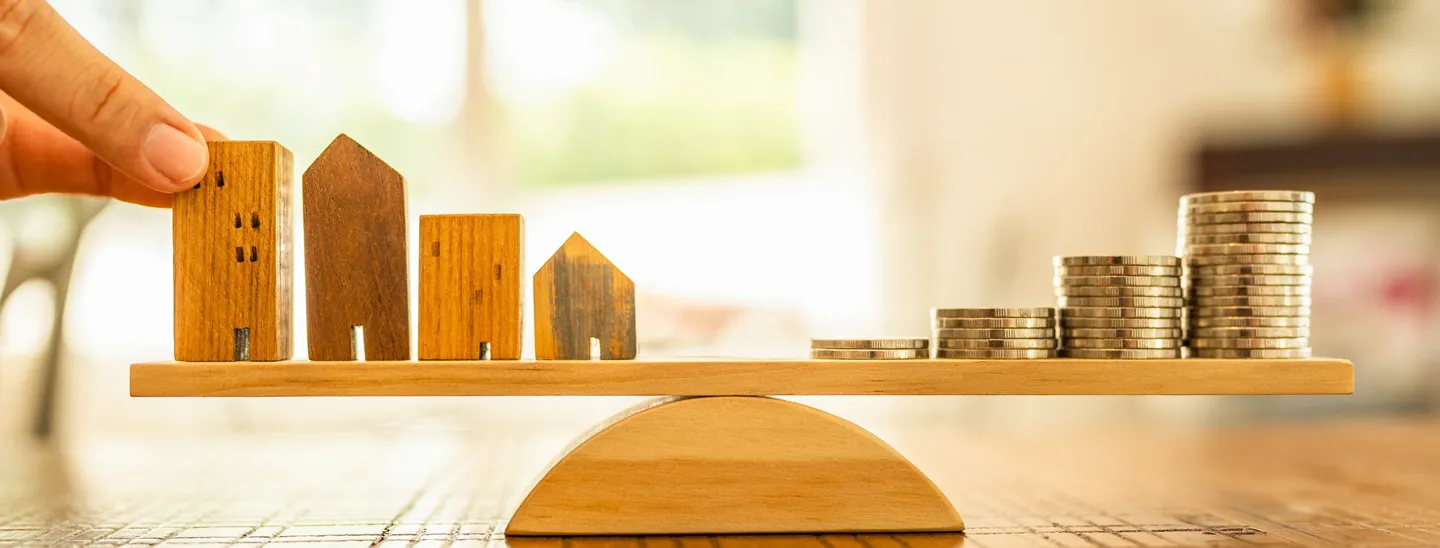 The image shows four wooden house models on one side of a wooden balance scale and stacks of coins on the other side. This visual represents the concept of weighing cash value against cash surrender value in life insurance planning
