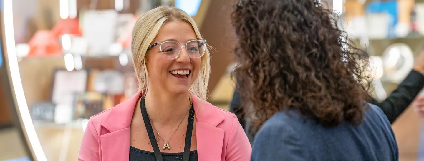 Two professionals engaged in conversation in a modern office setting, one wearing a pink blazer and the other in a dark jacket, representing pay and benefits discussions and workplace collaboration.
