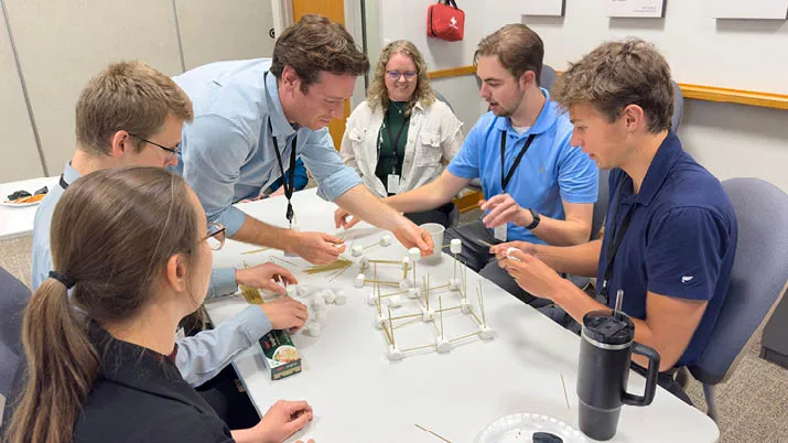 Interns participating in a hands-on team-building activity, constructing a structure with marshmallows and wooden sticks on a table in an office setting, representing collaboration and internship opportunities.
