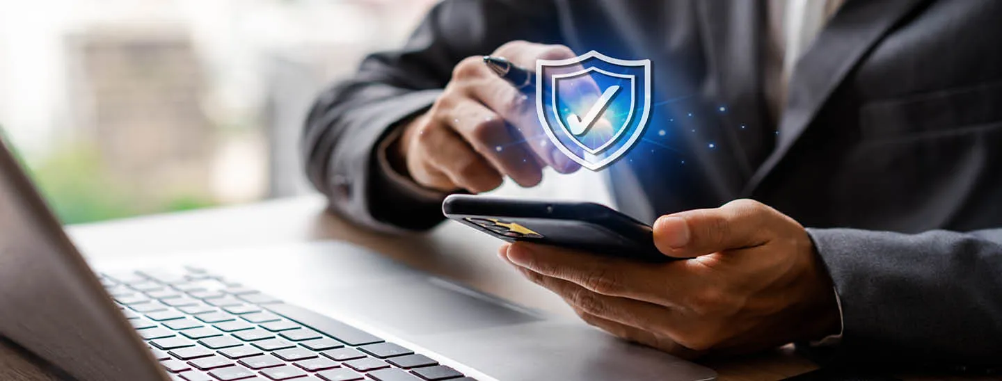 Employee using smartphone in front of a laptop performing a cybersecurity check