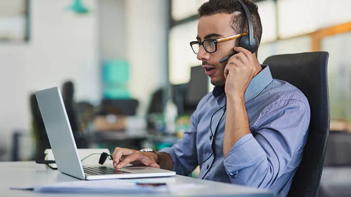 A professional seated at a modern office desk, using a laptop and wearing a headset for communication. The bright, open workspace reflects a collaborative and technology-focused environment, ideal for careers in customer service, IT support, and digital operations.