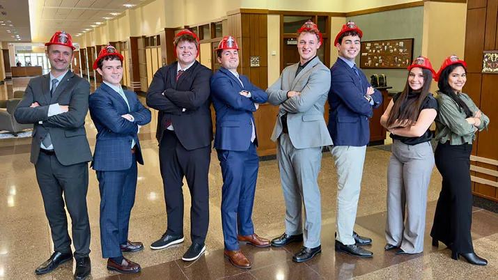 Group of interns standing in a polished office hallway wearing business attire and red plastic firefighter hats, representing internship opportunities, team spirit, and a fun workplace culture.