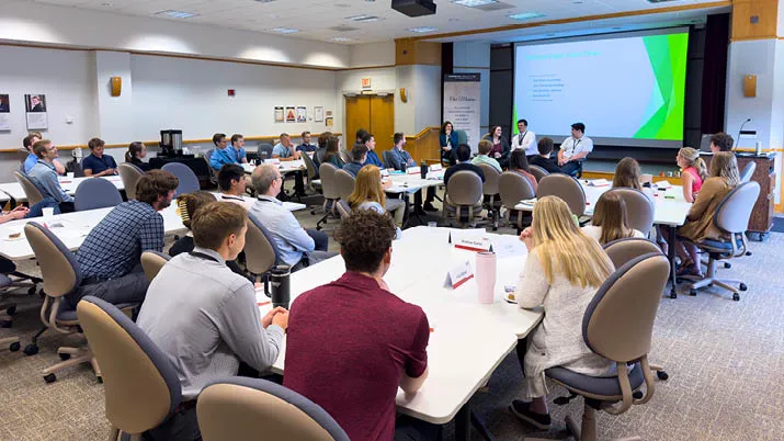 Internship group attending a professional development session in a conference room, seated at round tables facing a presentation screen displaying slides, representing learning, networking, and career growth opportunities.
